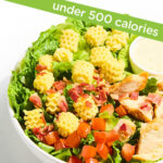A salad bowl with lettuce, diced tomatoes, bacon bits, grilled chicken pieces, crinkle-cut pasta, and a side of dressing. A label above reads “under 500 calories.