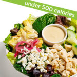 A salad bowl with leafy greens, diced chicken, crumbled feta, artichoke hearts, pickled red onions, cucumber slices, and a side of dressing. A label above reads “under 500 calories.”