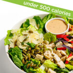 A salad bowl containing leafy greens, roasted Brussels sprouts, red quinoa, pickled red onions, grilled tofu cubes, crumbled feta, and a side of dressing. A label above reads “under 500 calories.”