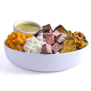 White bowl with steak cubes, roasted Brussels sprouts, sweet potato cubes, cottage cheese, and a small cup of green sauce on a light gray background.