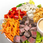 Salad bowl with steak cubes, shredded carrots, roasted red peppers, blue cheese crumbles, crispy onions, lettuce, and a cup of creamy dressing on a light gray background.