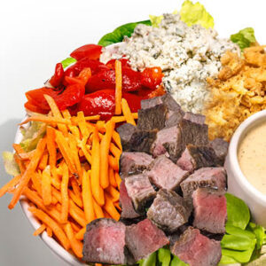 Salad bowl with steak cubes, shredded carrots, roasted red peppers, blue cheese crumbles, crispy onions, lettuce, and a cup of creamy dressing on a light gray background.